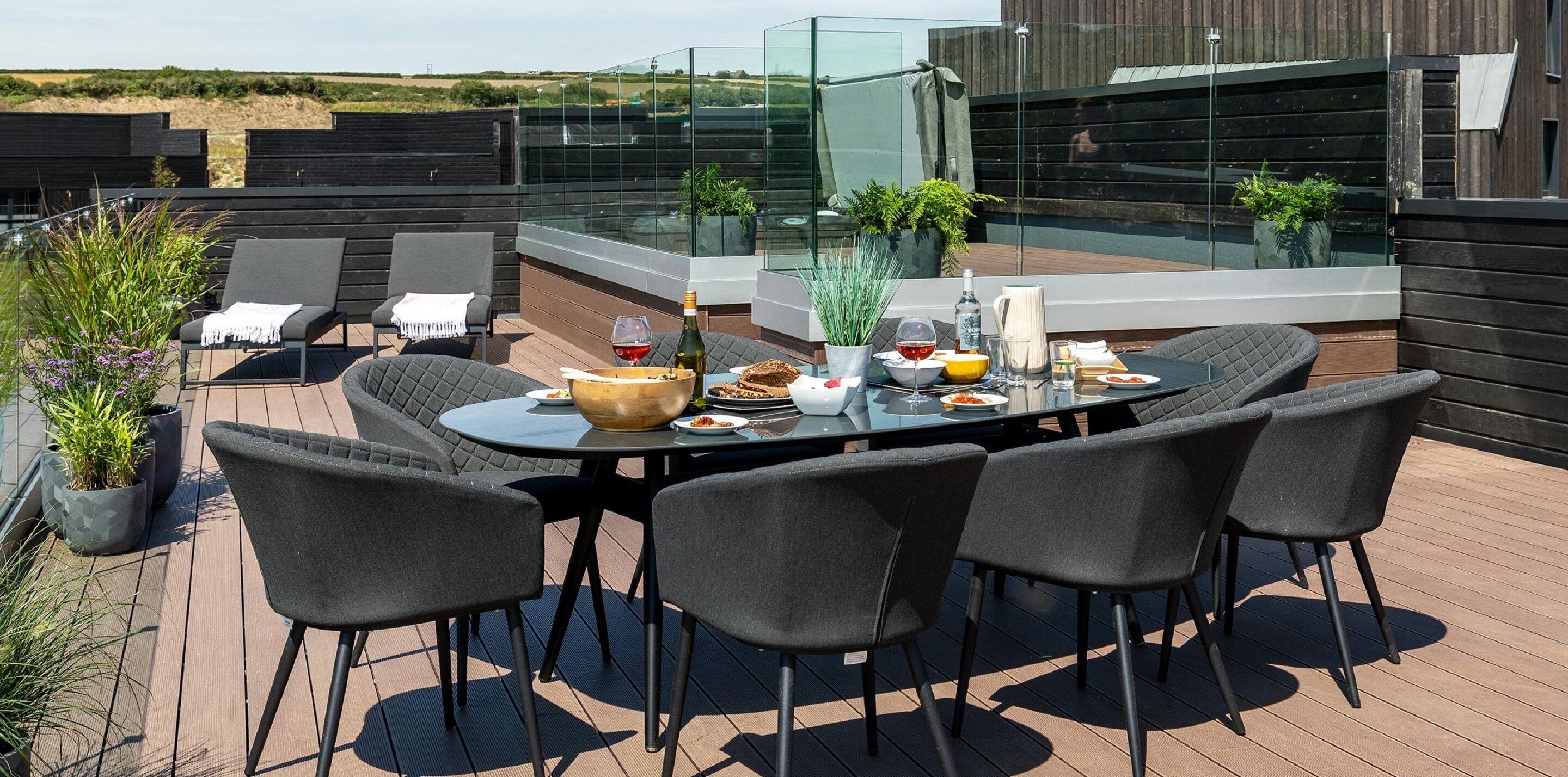 A rooftop terrace with a round table set for dining, surrounded by four chairs. There are potted plants and a glass railing.
