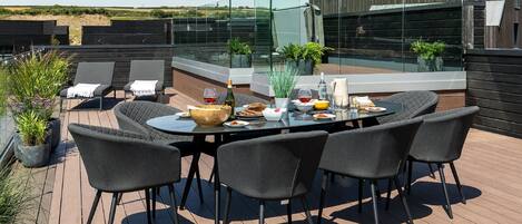 A rooftop terrace with a round table set for dining, surrounded by four chairs. There are potted plants and a glass railing.