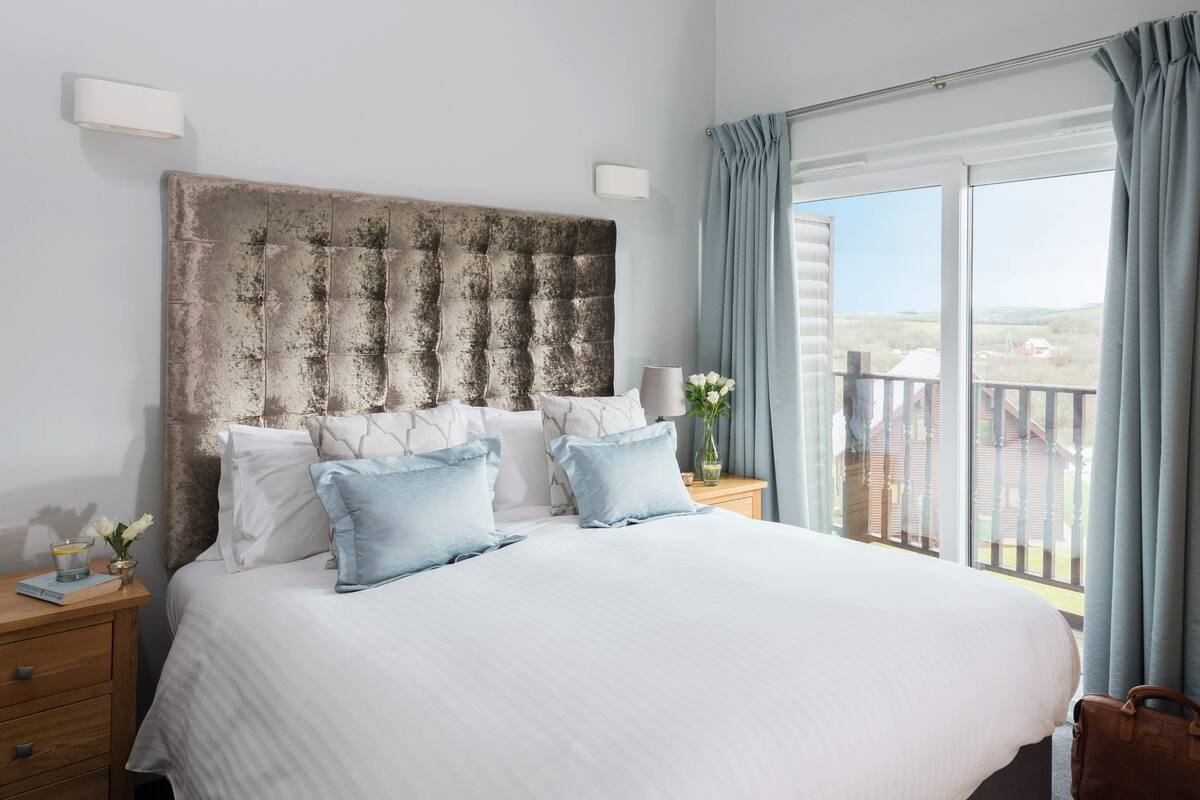 A large bed with a marble headboard, white bedding, and light blue pillows. There is a wooden nightstand with a lamp and a vase with flowers. A window with curtains and a view of buildings is visible.