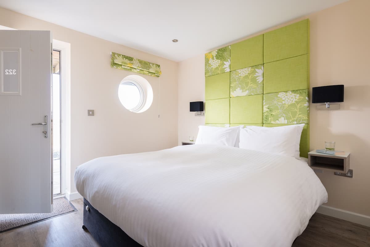 A bedroom with a large bed, a green headboard, two bedside tables with lamps, a round window, and a door with a number.
