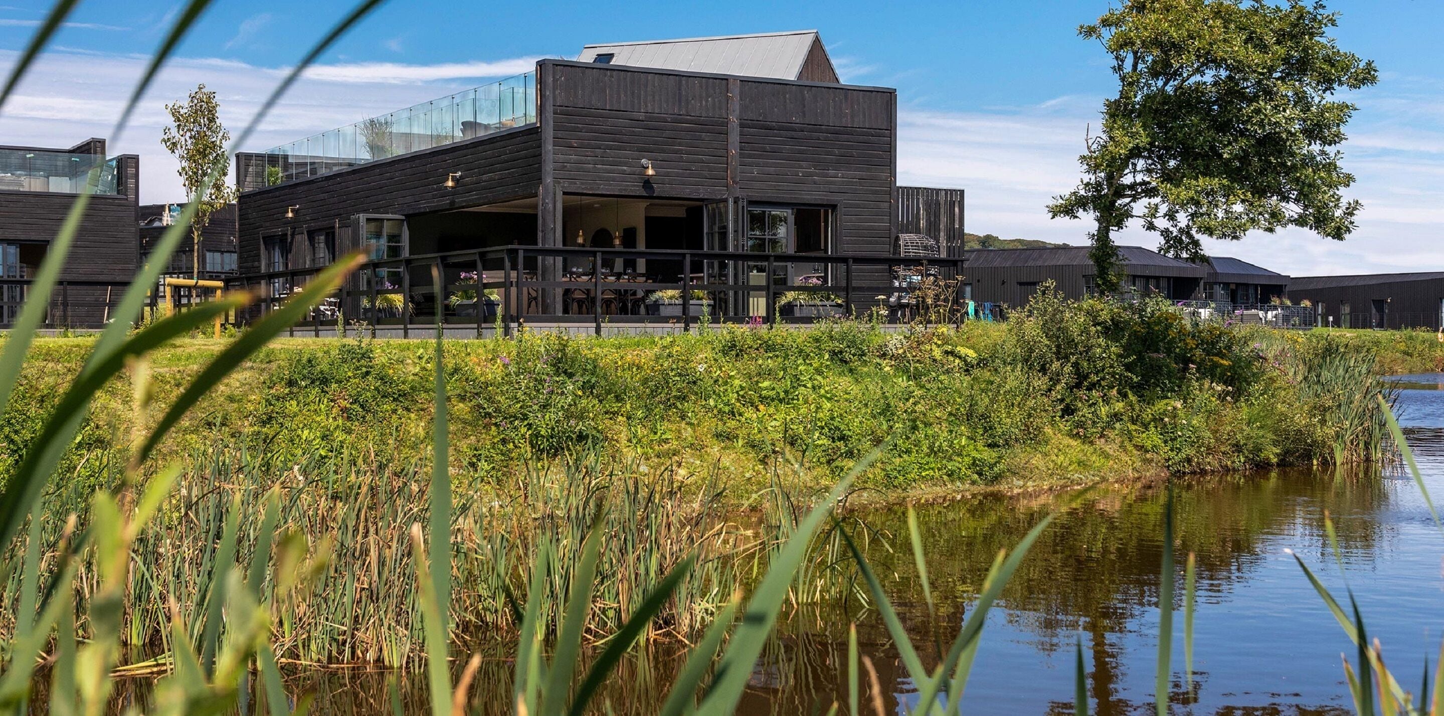 En moderne bygning med fladt tag og store vinduer, omgivet af grøn vegetation og et vandareal.