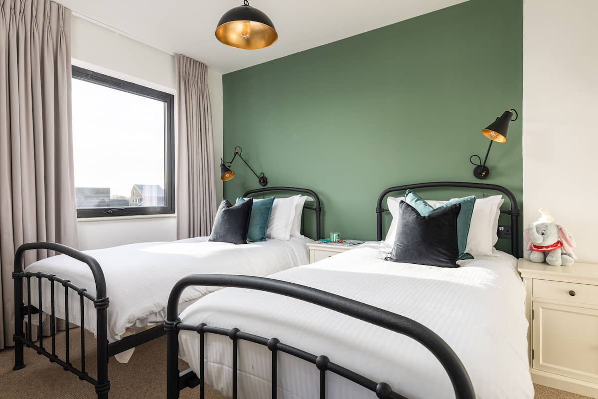 A twin bedroom with two beds, a window with curtains, a nightstand with a lamp, and a stuffed toy on the nightstand.