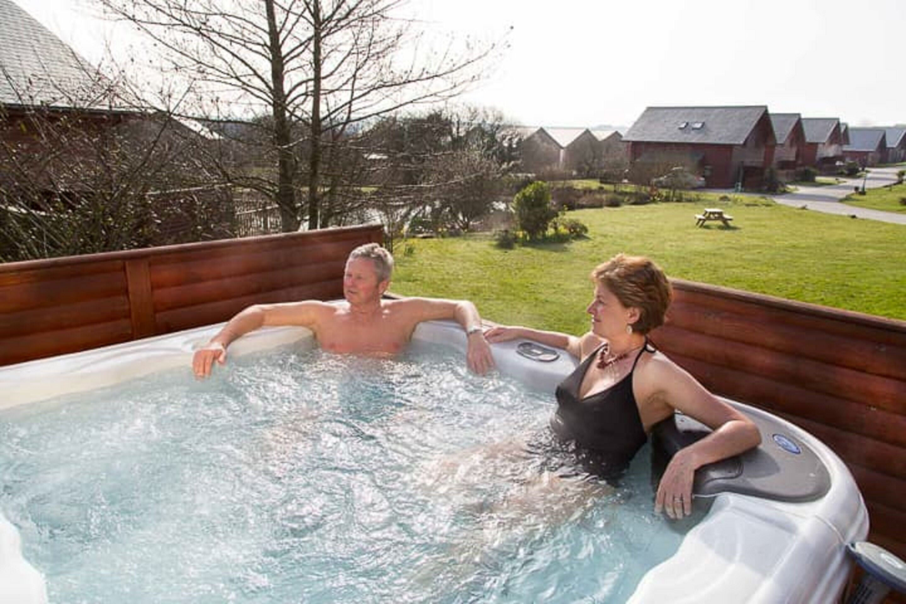 Two people in a hot tub outdoors with a grassy yard and houses in the background.