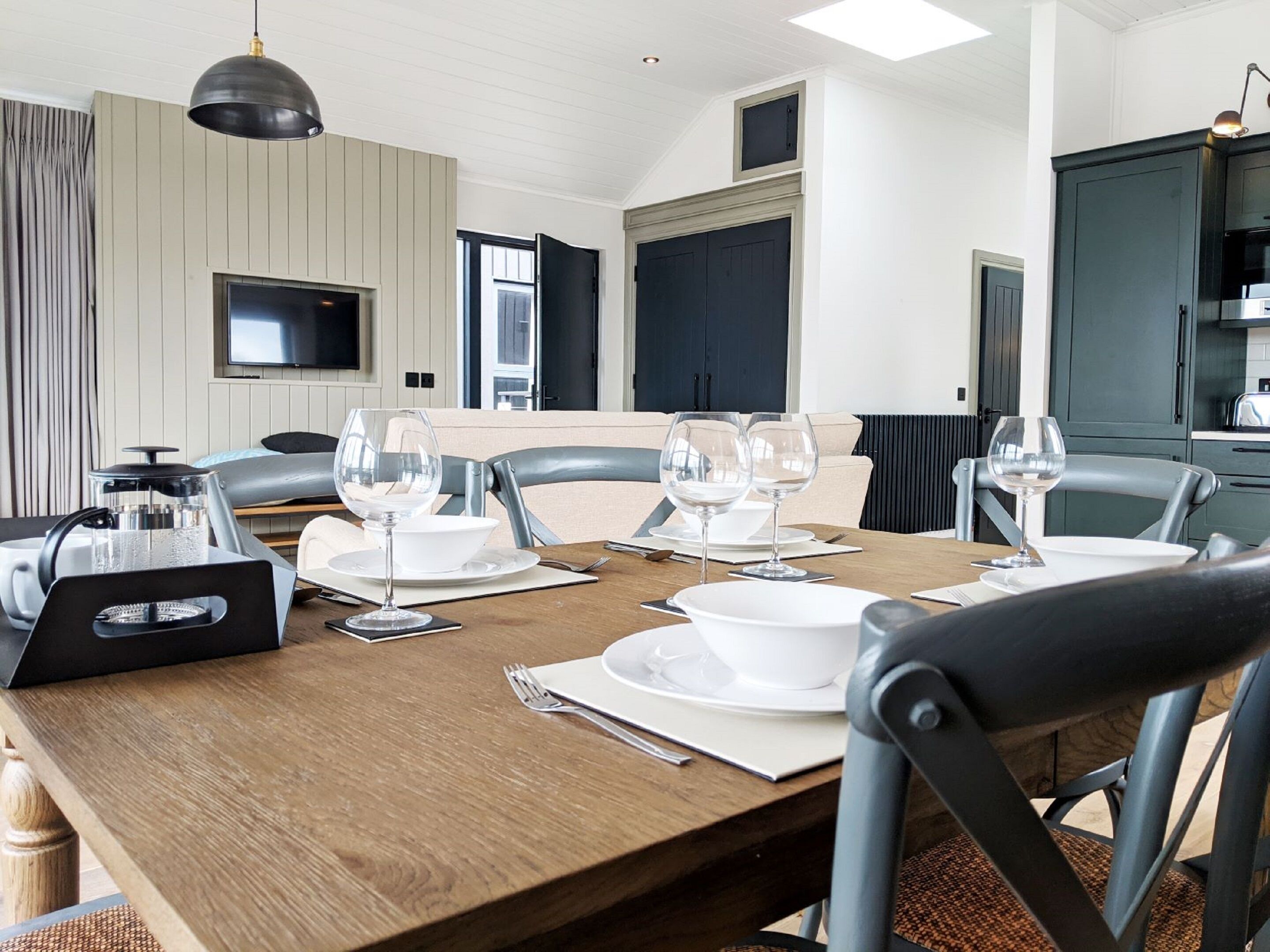 A dining area with a wooden table set for four, equipped with wine glasses, plates, and cutlery. A coffee maker and a kettle are on the table. The room has a modern design with a flat-screen TV mounted on the wall and a ceiling light fixture.