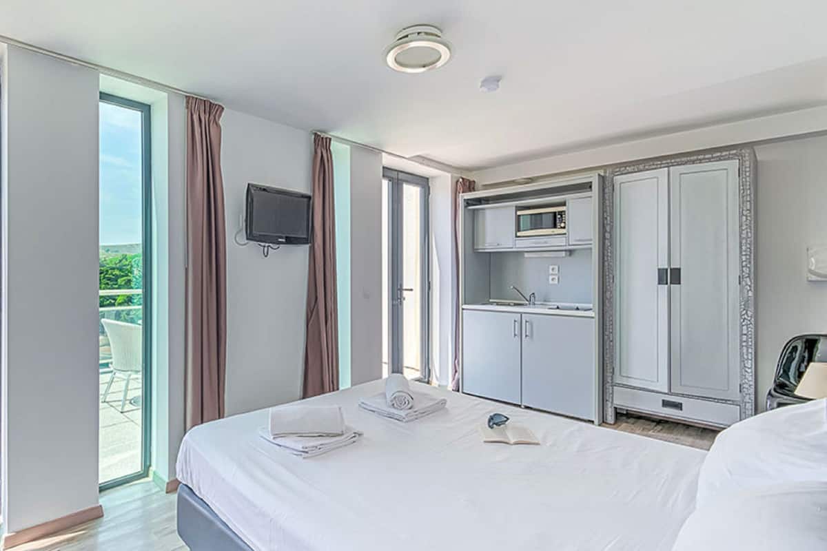 Ein Hotelzimmer mit Bett, Wandfernseher, Küchenzeile und Balkon mit Blick auf Grünflächen.