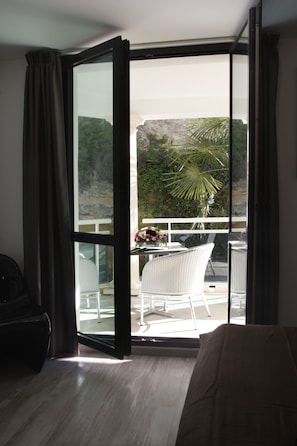 A room with a sliding glass door leading to an outdoor balcony with a white chair and a table, and a view of a rocky landscape.