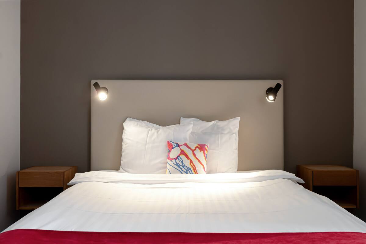 A neatly made bed with white linens, a colorful pillow, and two bedside tables with lamps.