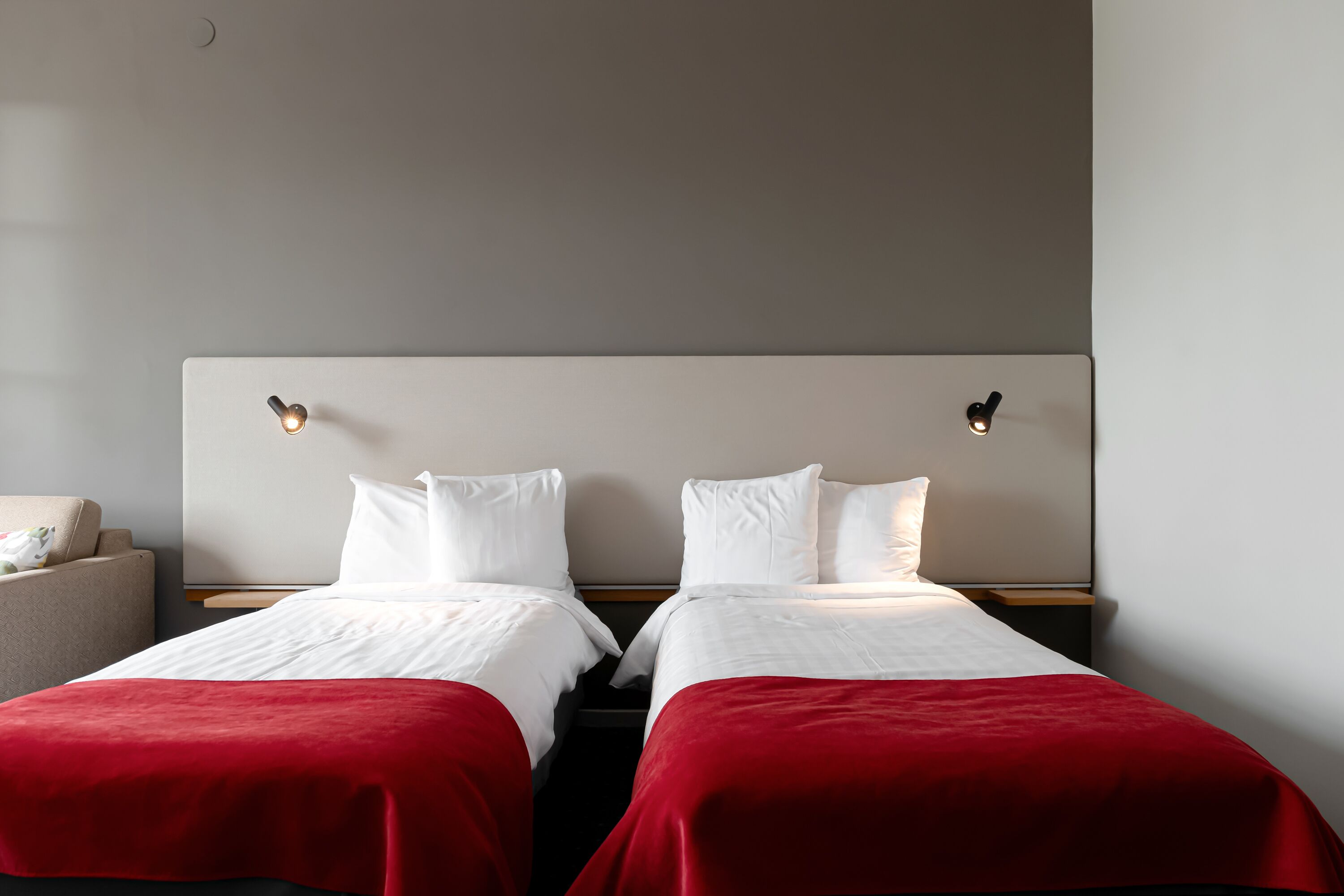 Habitación de hotel con dos camas, cada una con ropa blanca de cama y mantas rojas. Mesitas de noche con lámparas y una pequeña zona de estar con una silla y un cojín.