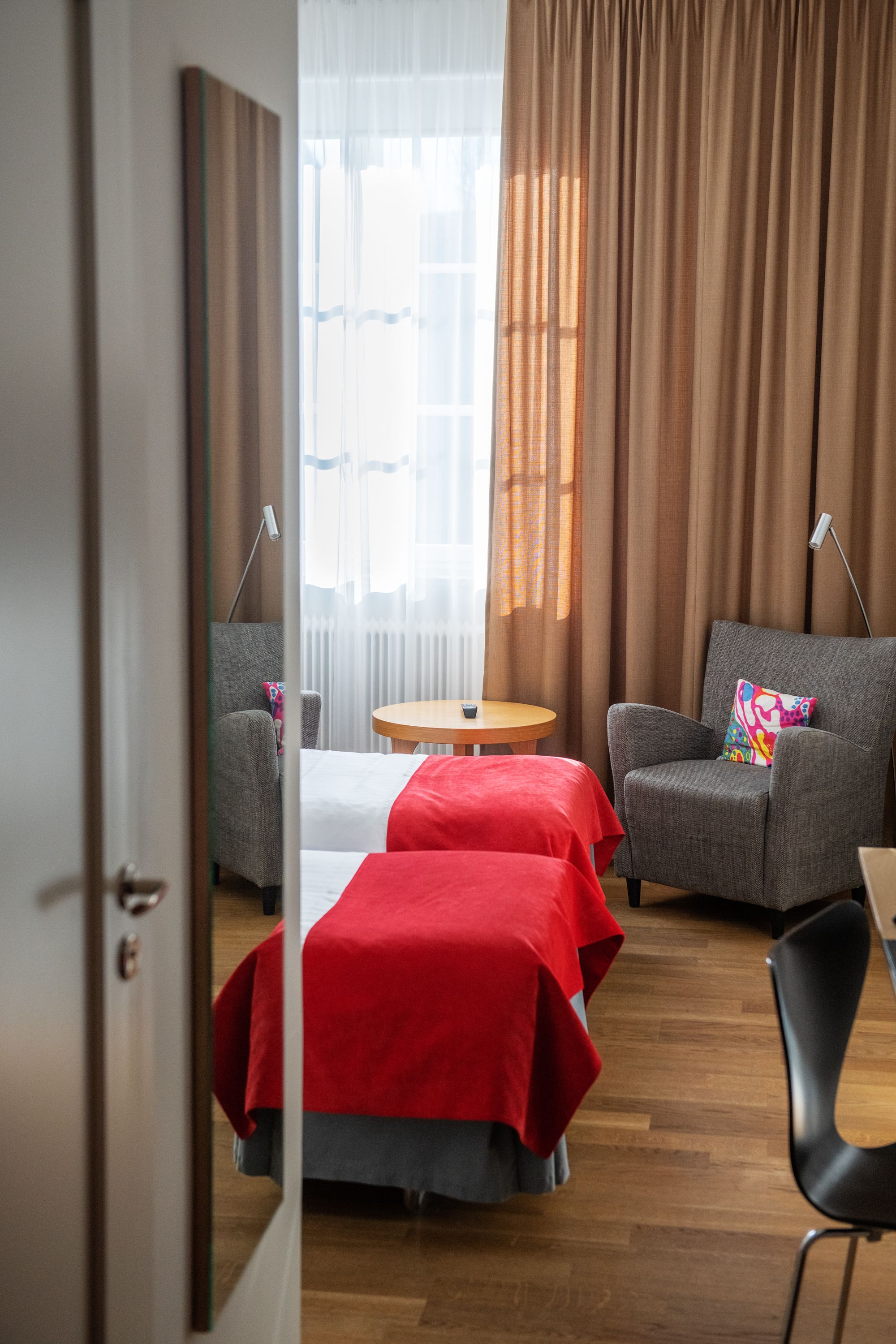 Habitación de hotel con cama, dos sillones, una mesita y vistas al exterior a través de cortinas traslúcidas.