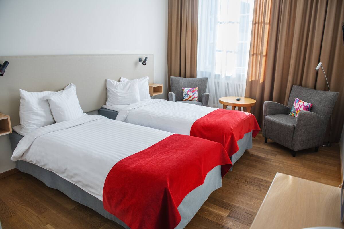A hotel room with two beds, a small table, a chair, and a view of the city through large windows.
