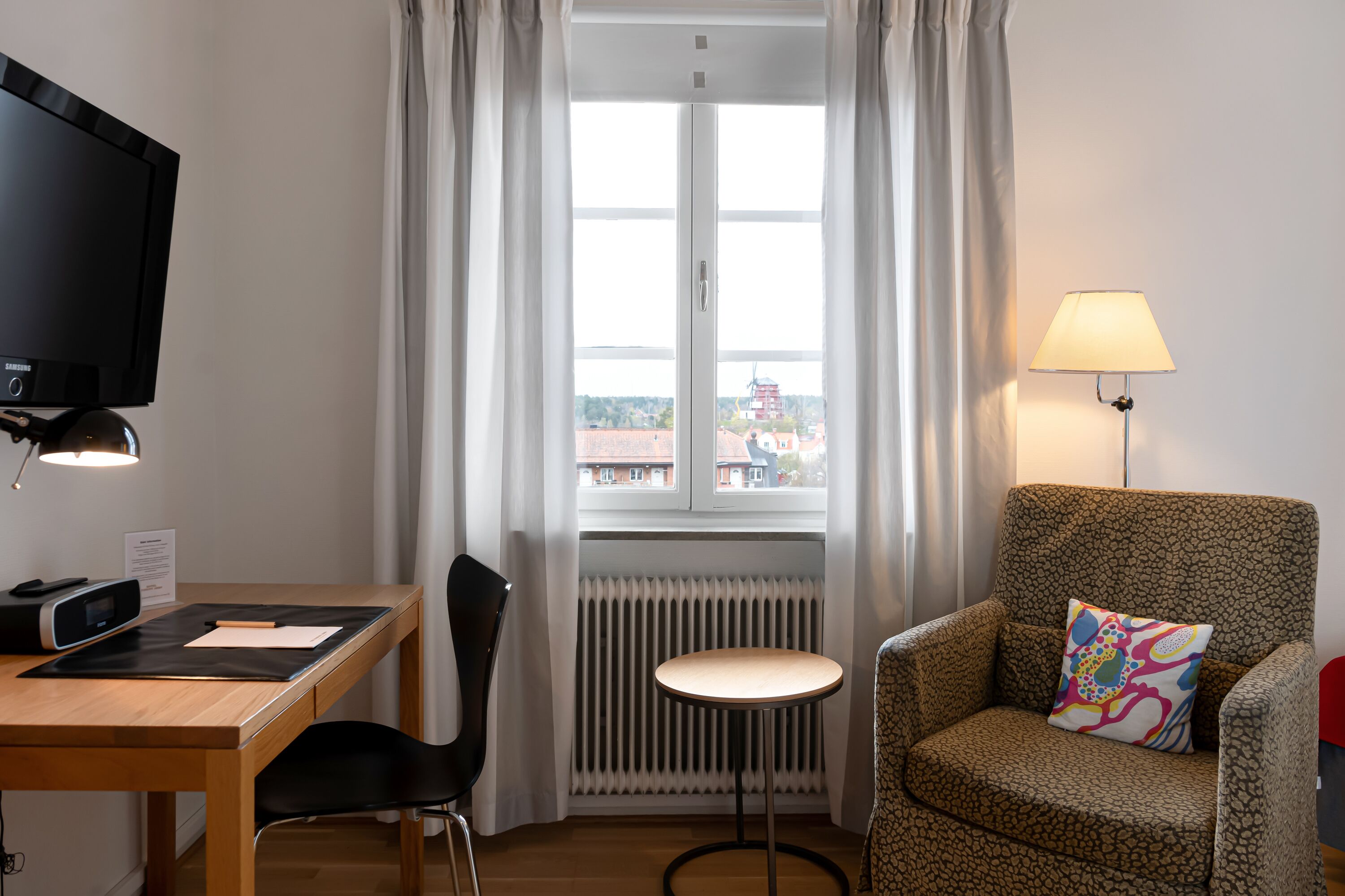 A hotel room with a desk, chair, TV, lamp, and a view of buildings through a window.