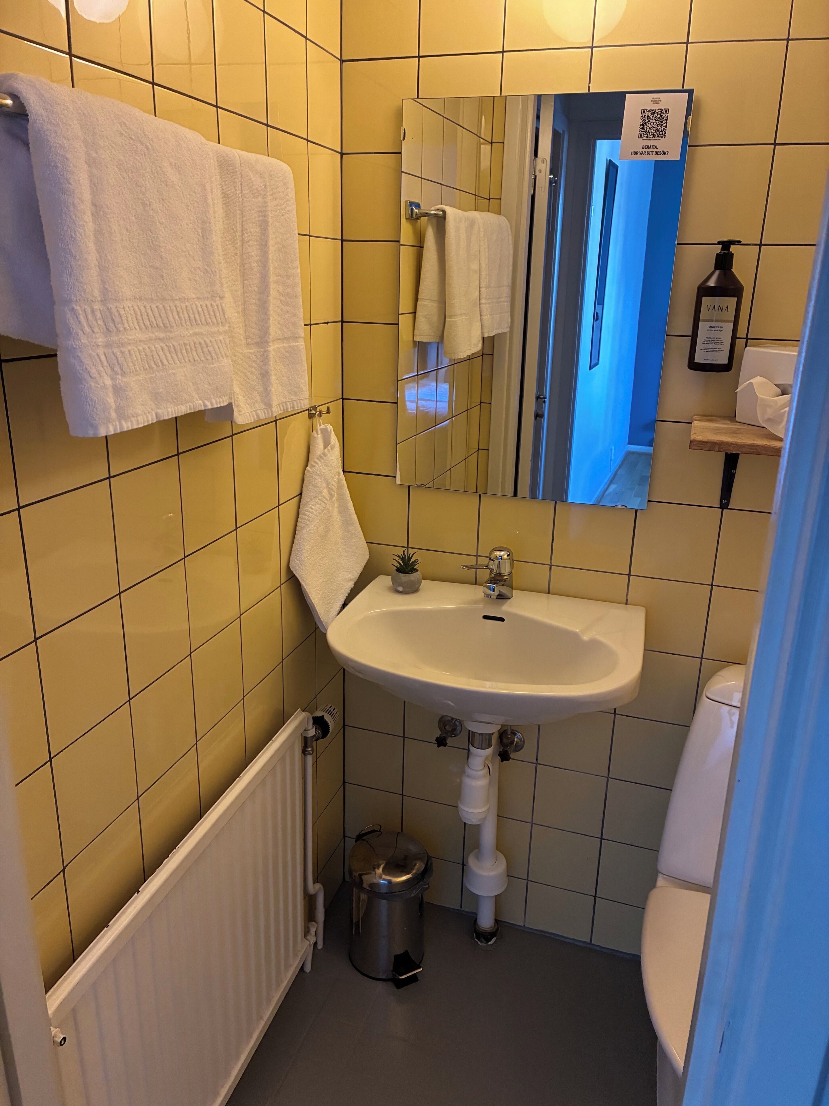 A bathroom with yellow tiles, a white sink, a mirror, a towel rack, a small trash can, and a toilet.