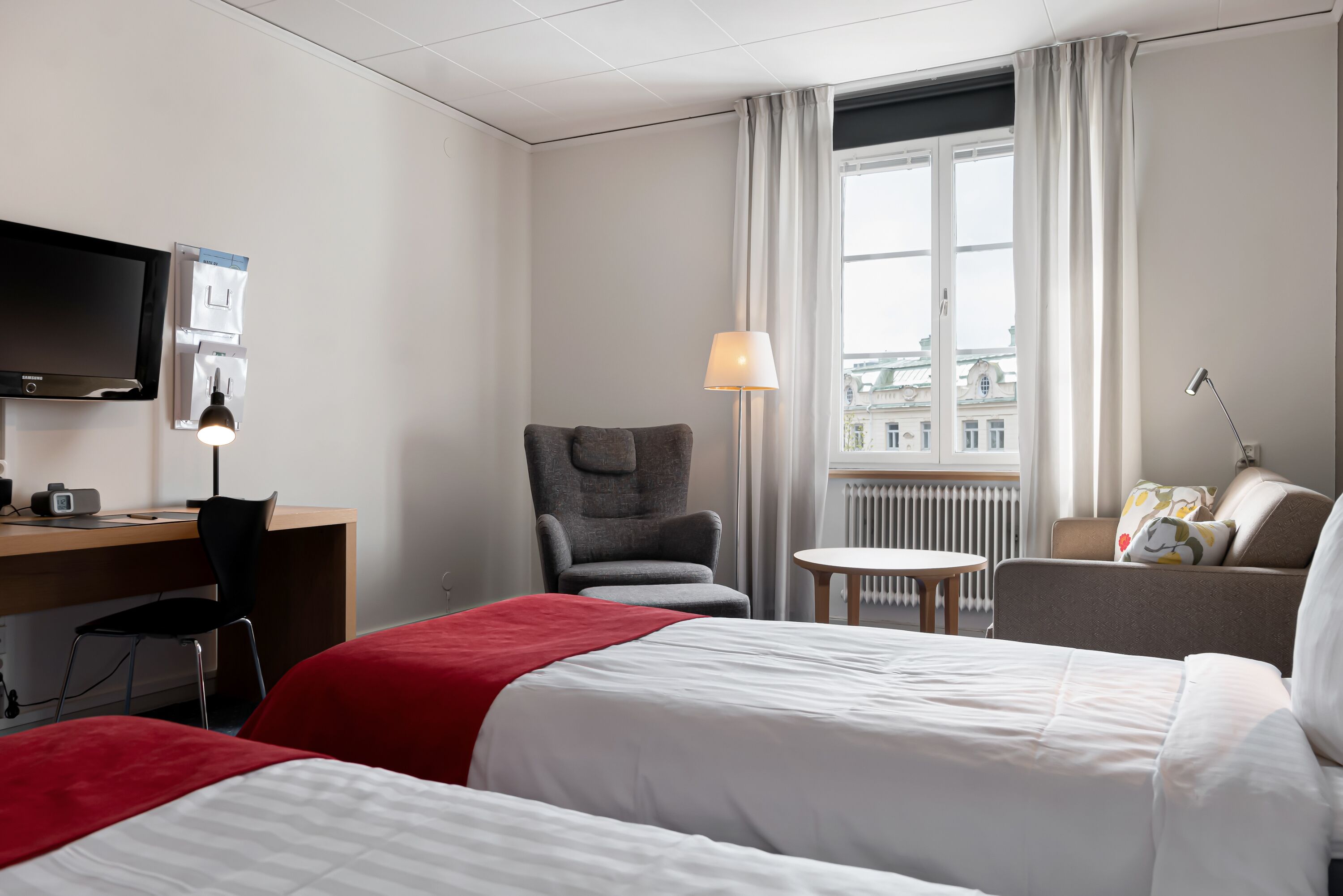 A hotel room with two beds, a desk with a chair, a TV, a lamp, and a view of buildings through the window.