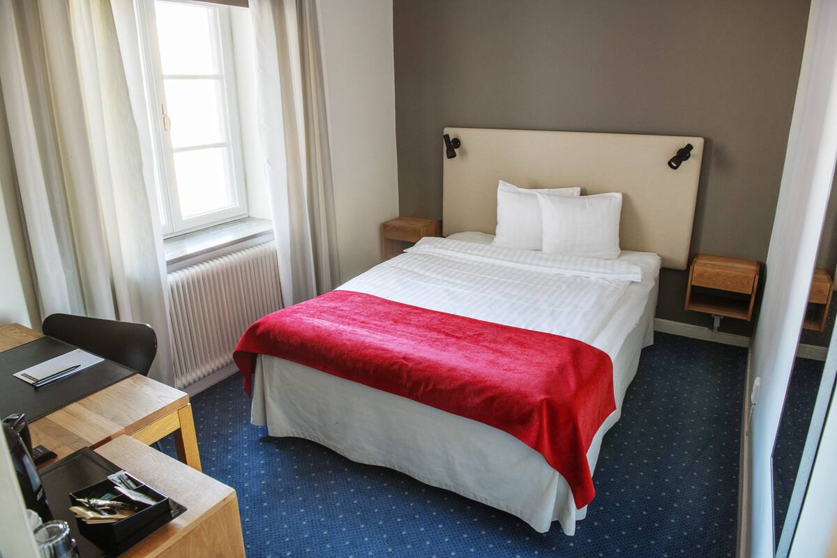 A hotel room with a bed, a desk, a chair, a window, and a radiator.