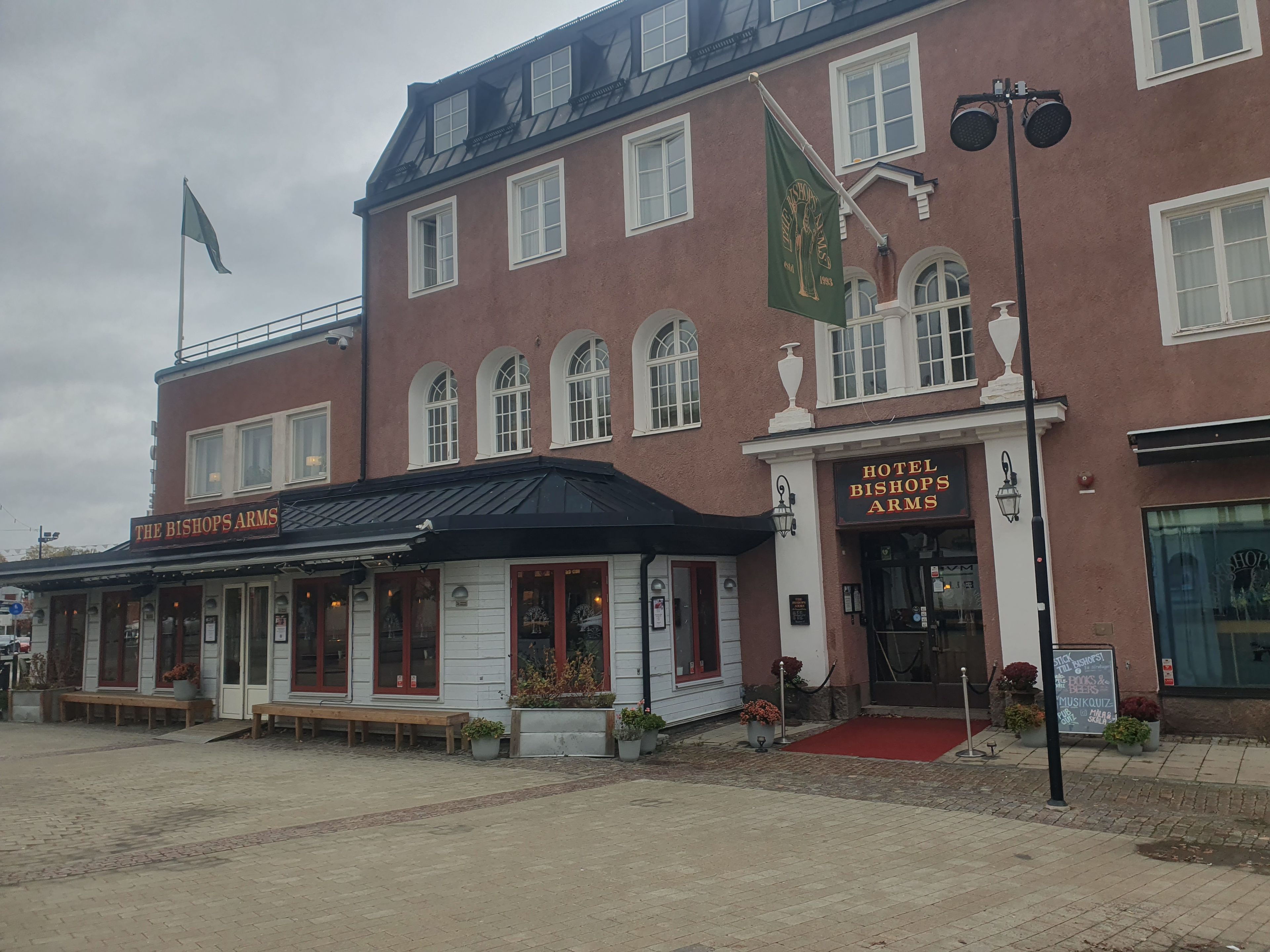 A building with a sign for 'The Bishops Arms' and 'Hotel Bishops Arms' indicating a hotel. There are benches outside and a red carpet leading to the entrance.