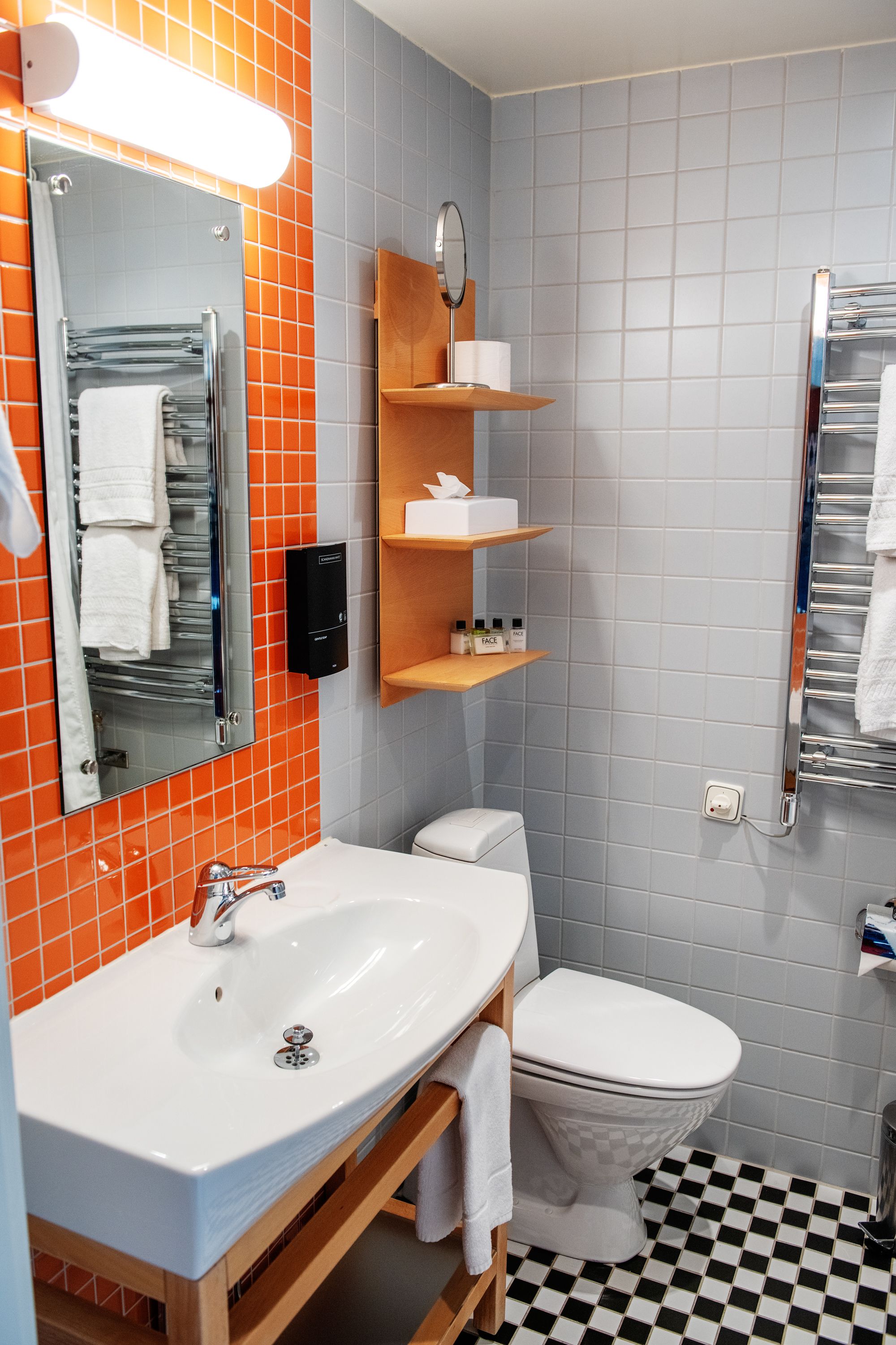 Un baño con lavamanos blanco, espejo y retrete. Las paredes tienen azulejos naranja y gris, y el piso es de cuadros negros y blancos.