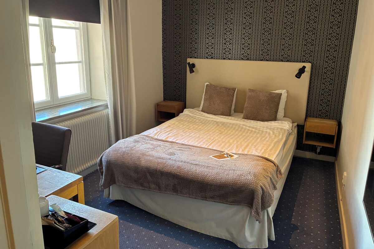 Een hotelkamer met een bed, een bureau, een stoel, een kleine tafel, een radiator en een behang met patroon.
