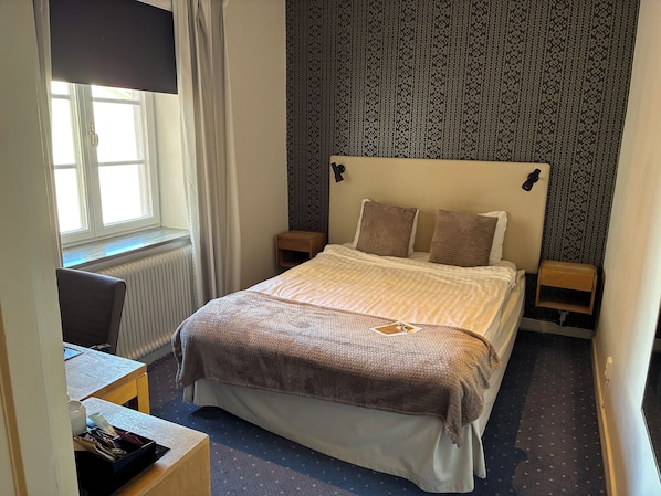 Habitación de hotel con cama, escritorio, silla, mesita pequeña, radiador y pared con estampado.