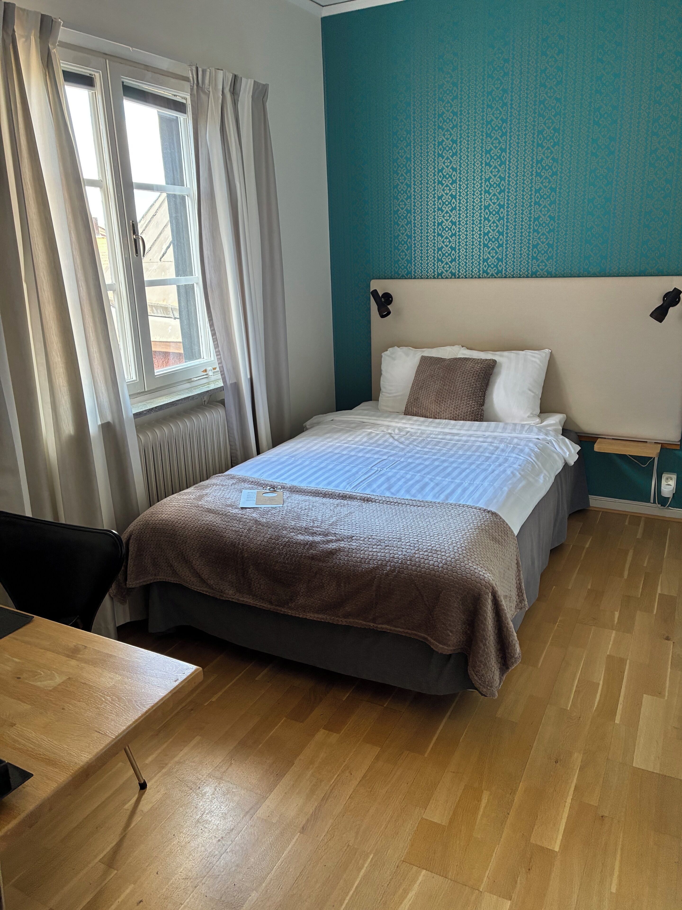 Habitació d'hotel amb un llit, una taula de treball, una finestra amb cortines i una paret de color verd marí.