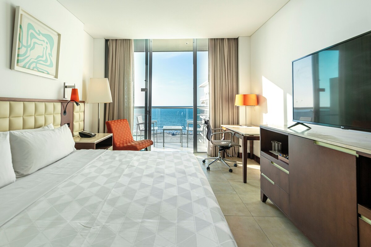 A hotel room with a large bed, a desk, a chair, a TV, and a view of the ocean.