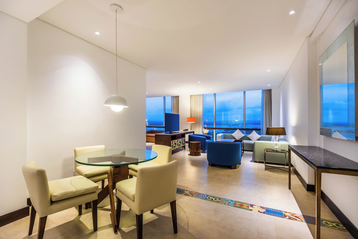 A modern hotel room with a dining area, seating, a TV, and large windows.