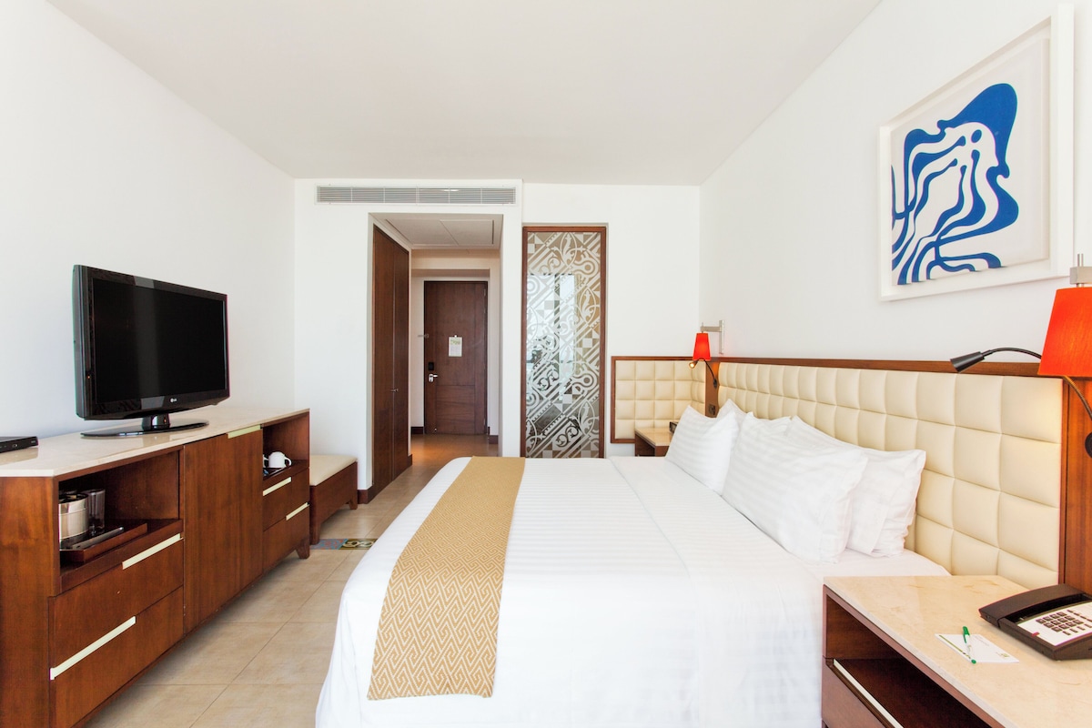 A hotel room with a bed, a television, a desk with a lamp, and a painting on the wall.