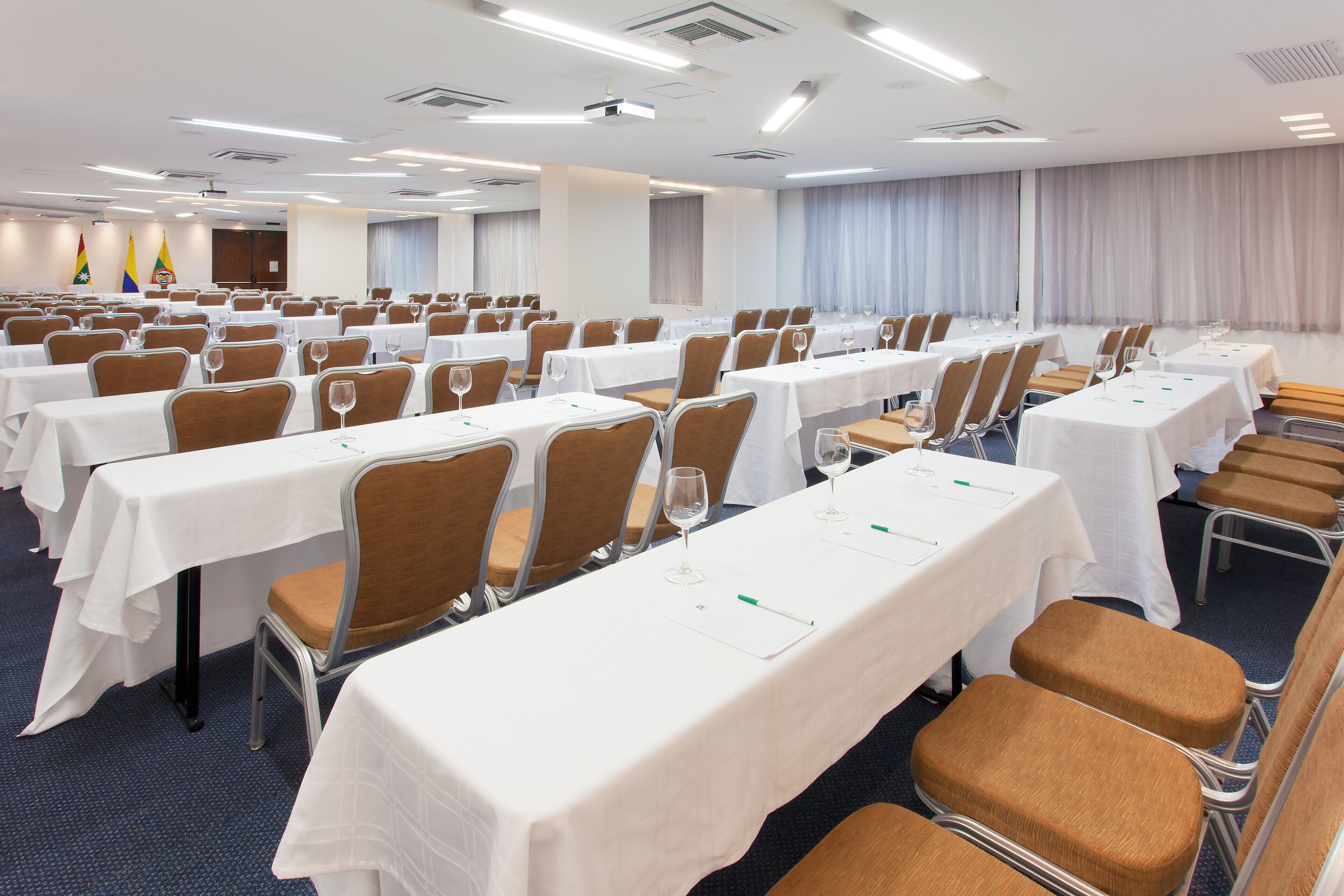 Uma sala de conferências com fileiras de mesas e cadeiras, toalhas de mesa brancas e bandeiras ao fundo.