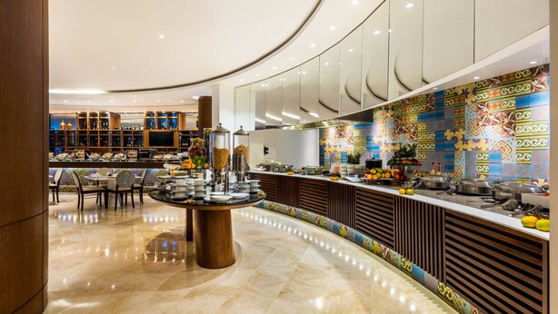 Eine moderne Hotelhalle mit einem runden Tisch in der Mitte, einem Buffetbereich mit verschiedenen Speisen und einer Wand mit geometrischen Mustern.