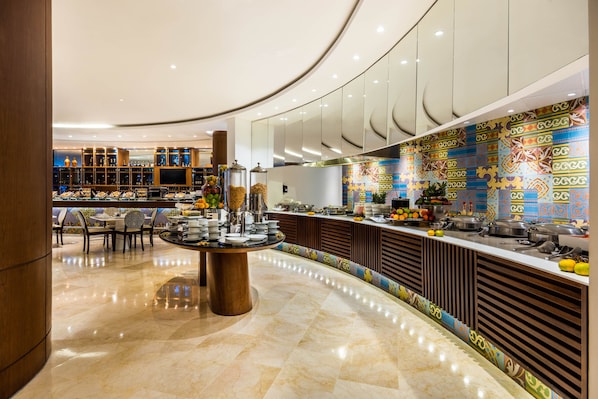 A modern hotel lobby with a central round table, a buffet area with various food items, and a decorative wall with geometric patterns.