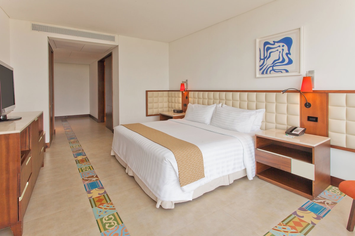 A hotel room with a bed, bedside tables, a desk, a television, and a hallway.