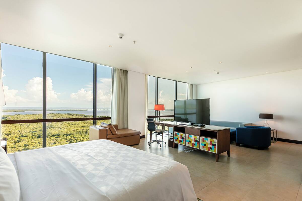 A hotel room with a large bed, a desk with a TV, a chair, and a view of a forested area through large windows.