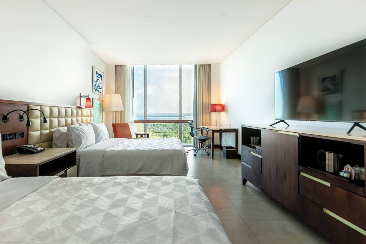 A hotel room with two beds, a desk, a TV, and a view of the outdoors.