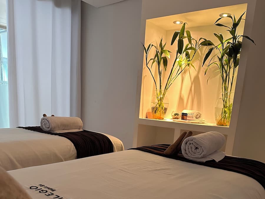 A hotel room with two beds, each with a rolled towel and a pillow. There is a wall-mounted shelf with plants and a small table with toiletries.