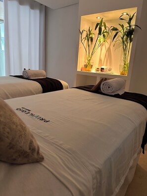 A hotel room with two beds, each with a rolled towel and a pillow. There is a wall-mounted shelf with plants and a small table with toiletries. - AC Hotel by Marriott Cartagena Colombia (Cartagena)