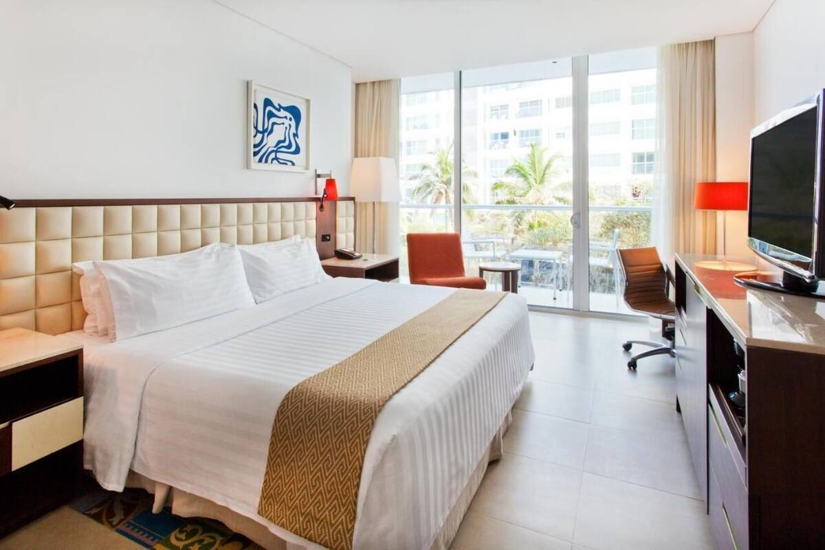 A hotel room with a large bed, a desk with a TV, a chair, and a balcony with a view of palm trees.