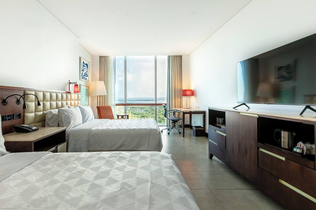A hotel room with two beds, a desk, a TV, and a view of the outdoors.