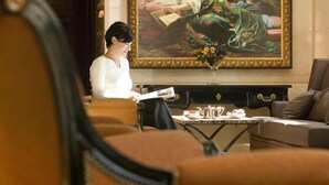 Une personne est assise dans une pièce, en train de lire. On y voit un grand tableau, une table avec un vase et un canapé.