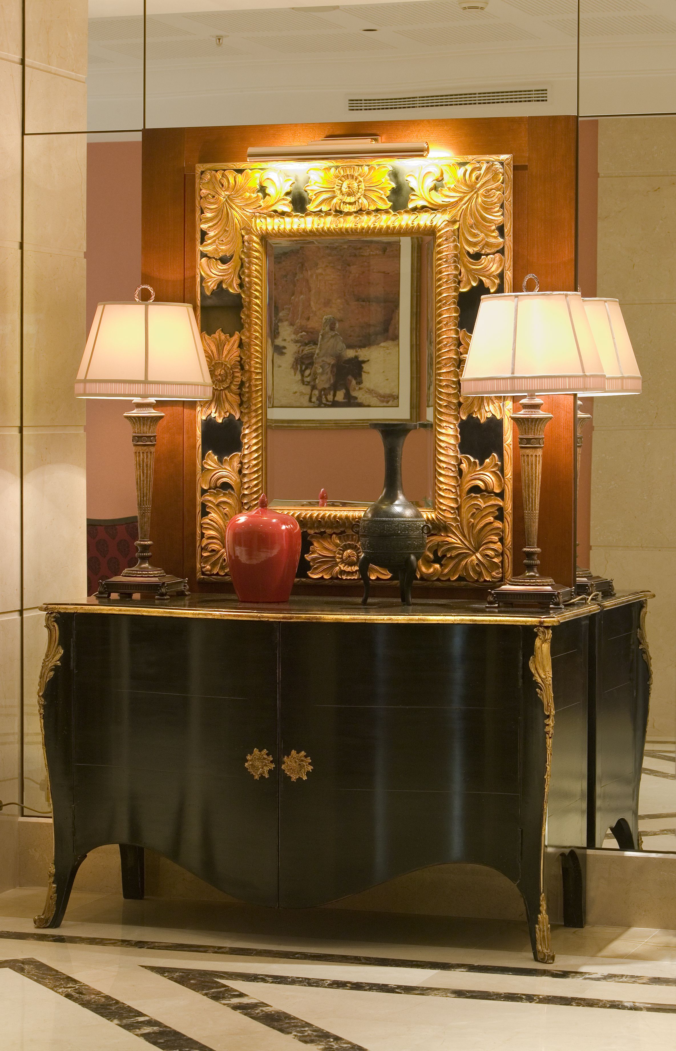 Una consola lujosa con dos lámparas, un florero rojo y un espejo con un marco ornamentado.