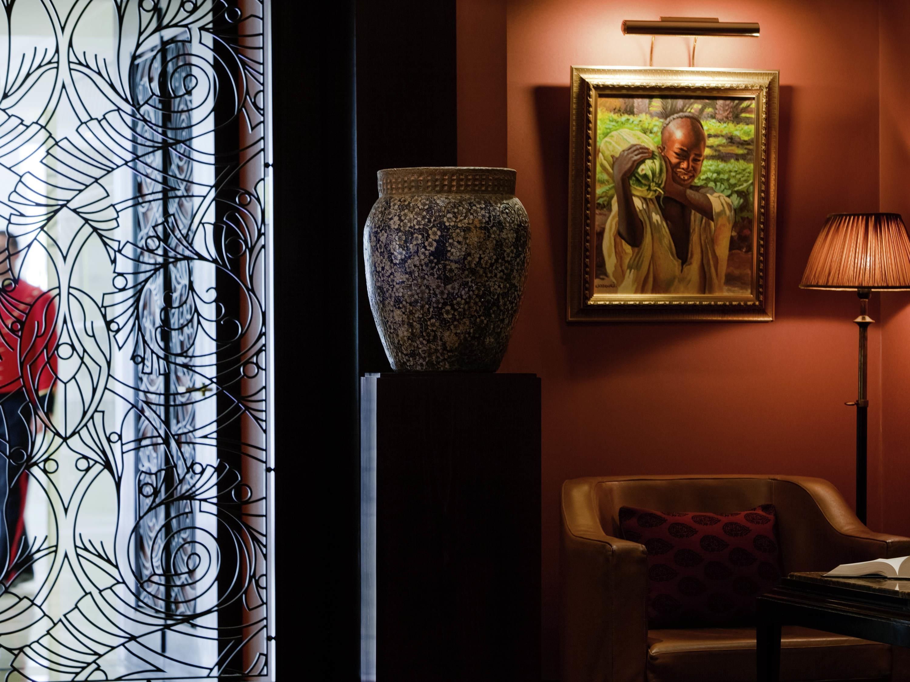 A room with a decorative metal gate, a large vase on a pedestal, a framed painting, a lamp, and a leather armchair.