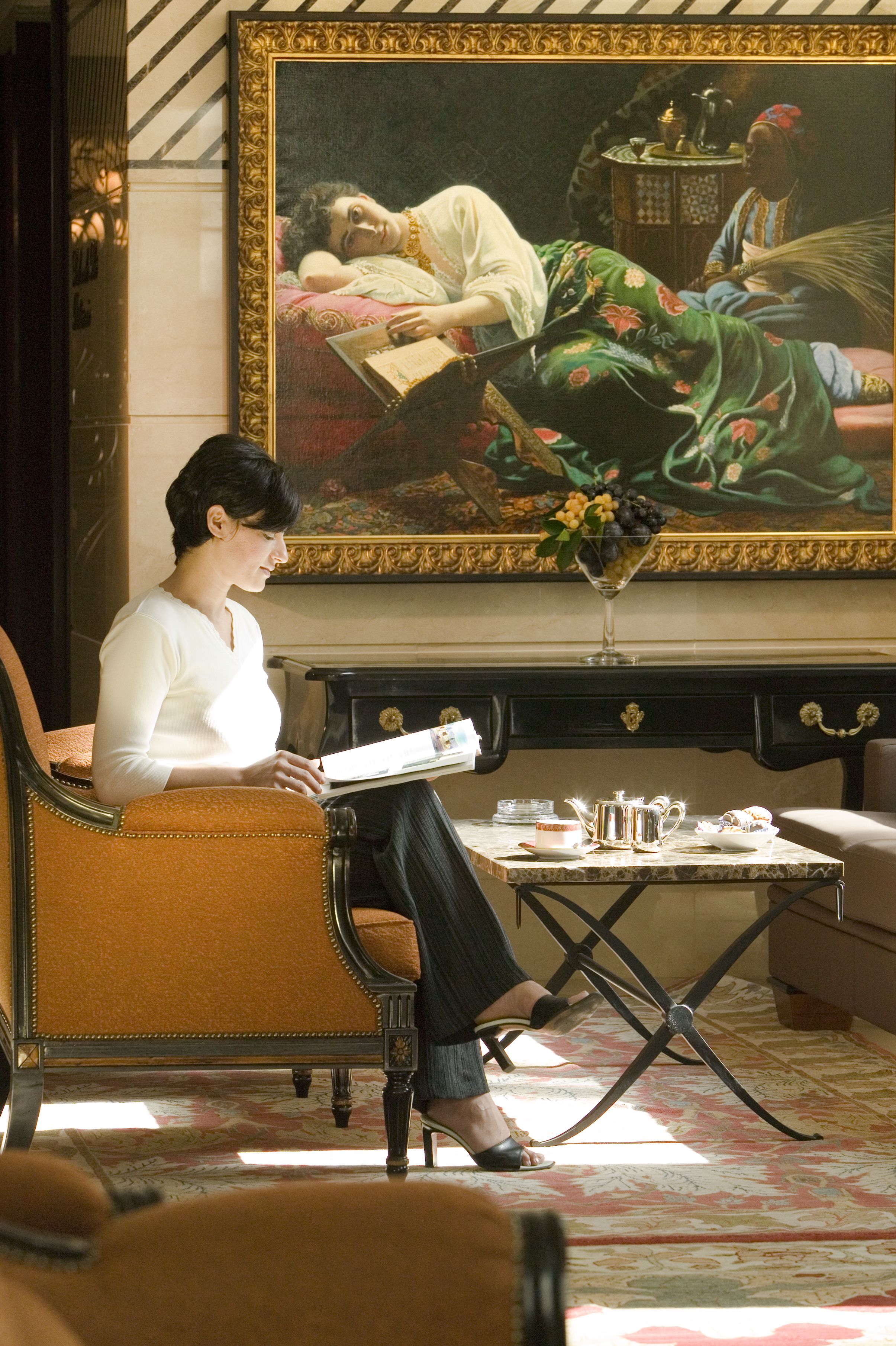 A woman seated in a room with a large painting on the wall, a table with a teapot and cups, and a patterned carpet.