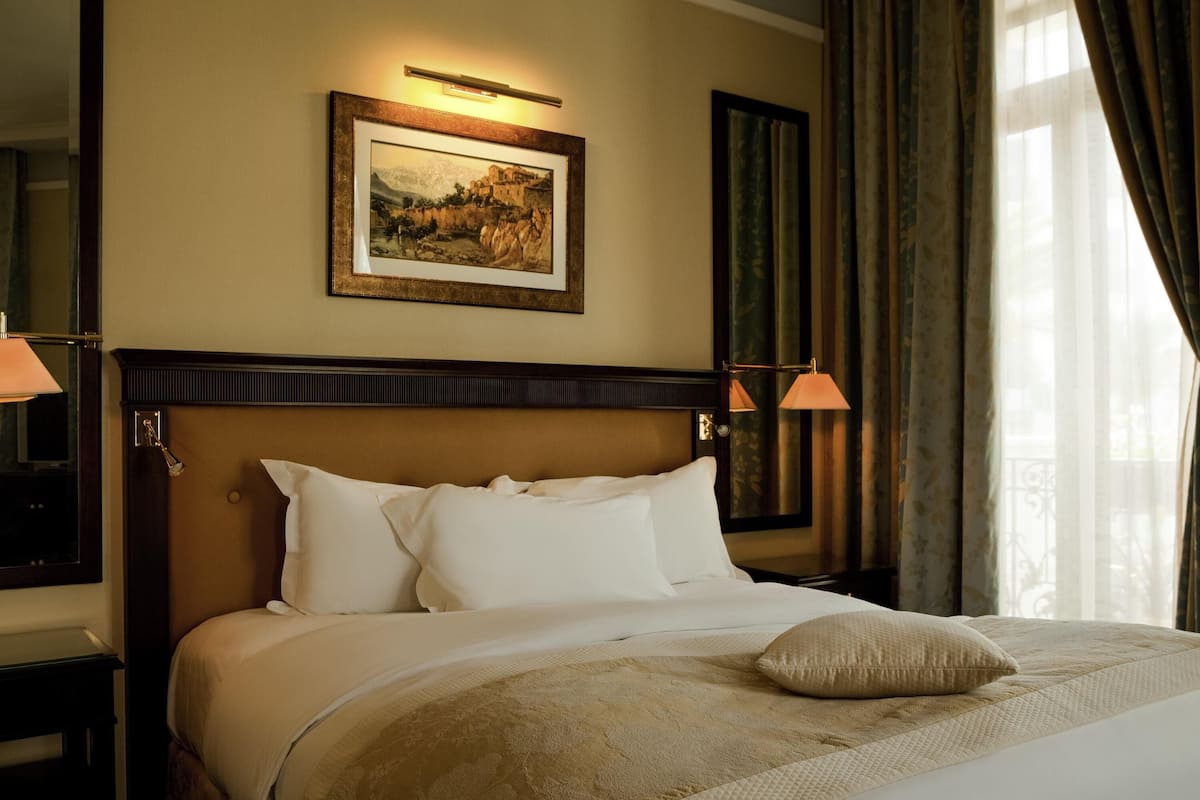 Une chambre d’hôtel avec un grand lit, une tête de lit ornée d’un tableau encadré, deux lampes de chevet et une fenêtre avec des rideaux.