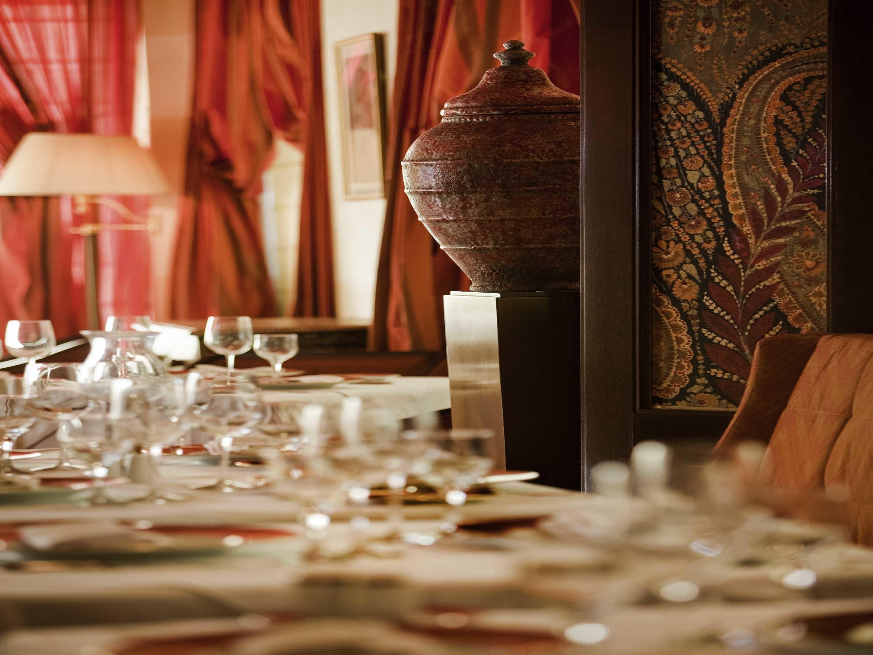 Une table à manger dressée avec de nombreux verres à vin et des assiettes, un grand vase décoratif et un mur orné d’un motif en arrière-plan.