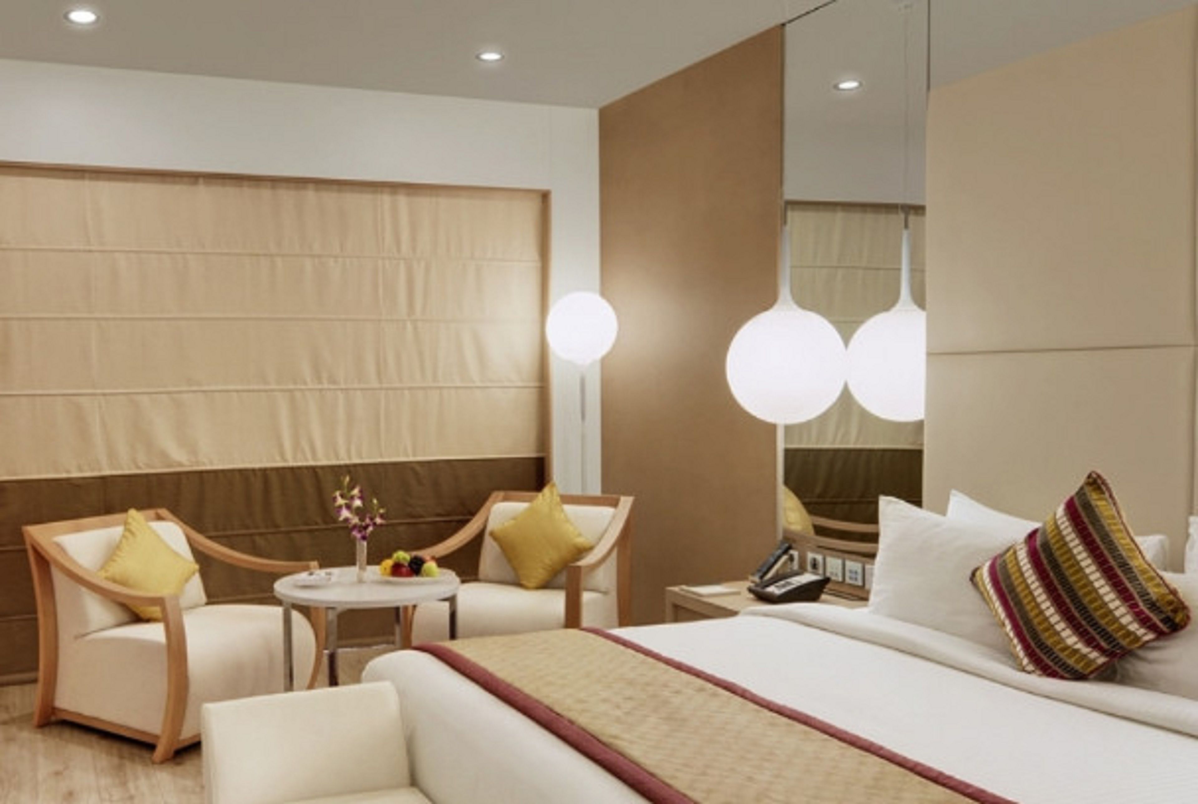 A modern hotel room with a large bed, two armchairs, a small round table with a vase of flowers, and a bedside table with a lamp and phone.