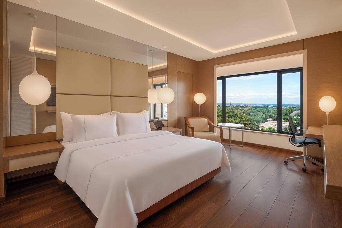 A modern hotel room with a large bed, a desk, a chair, and a view of the city through large windows.