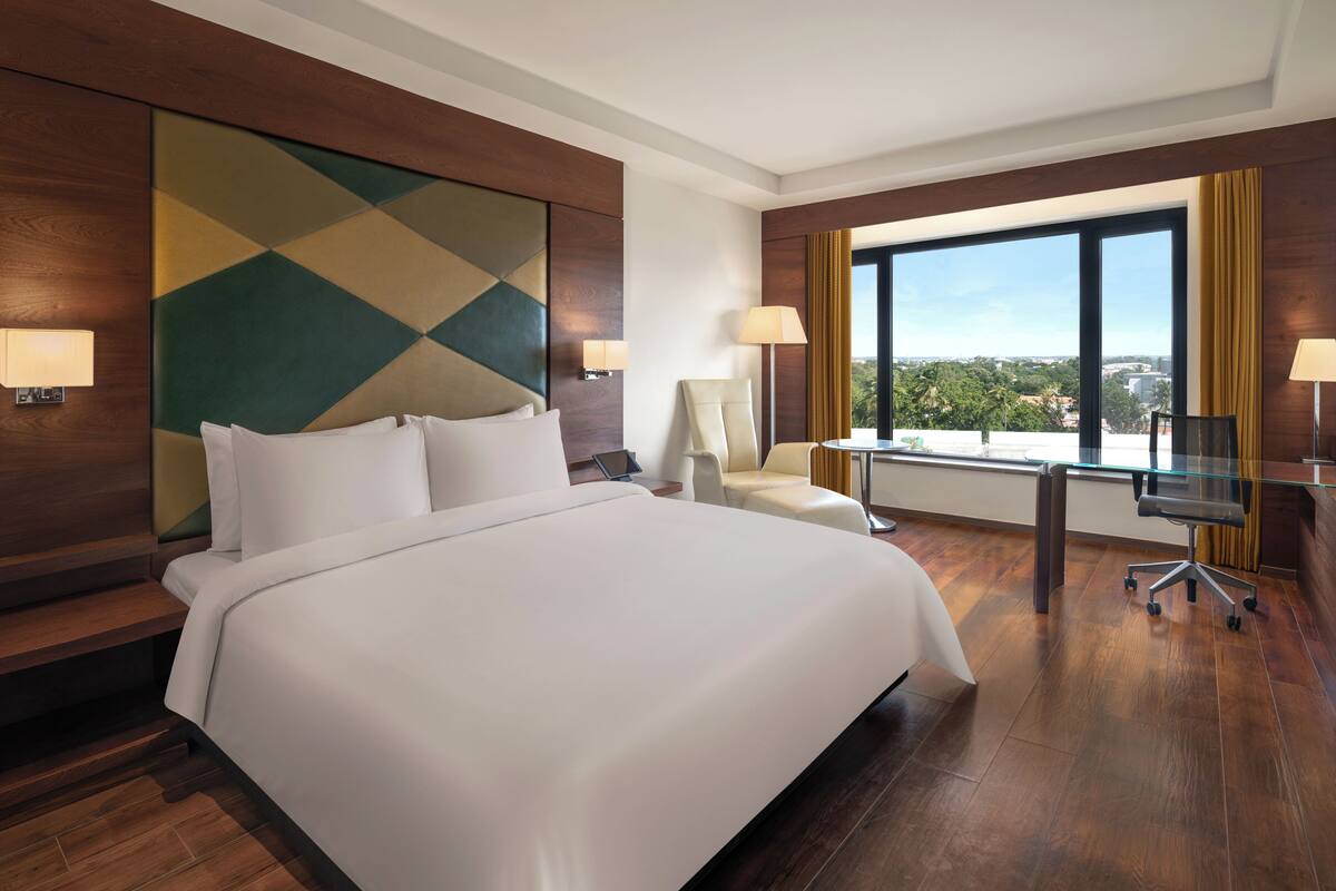 A hotel room with a large bed, a desk with a chair, a laptop, and a view of the city through large windows.