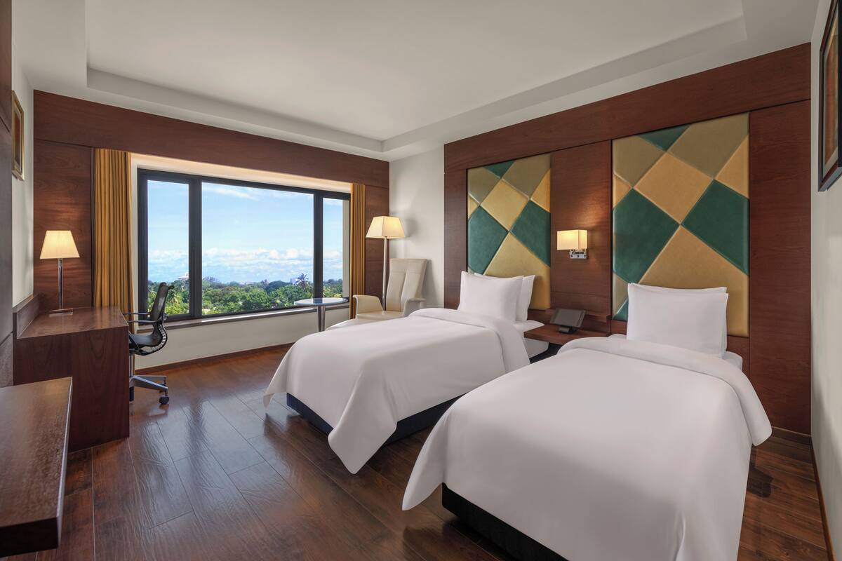 A hotel room with two beds, a desk, and a chair. There is a large window providing a view of the outdoors.