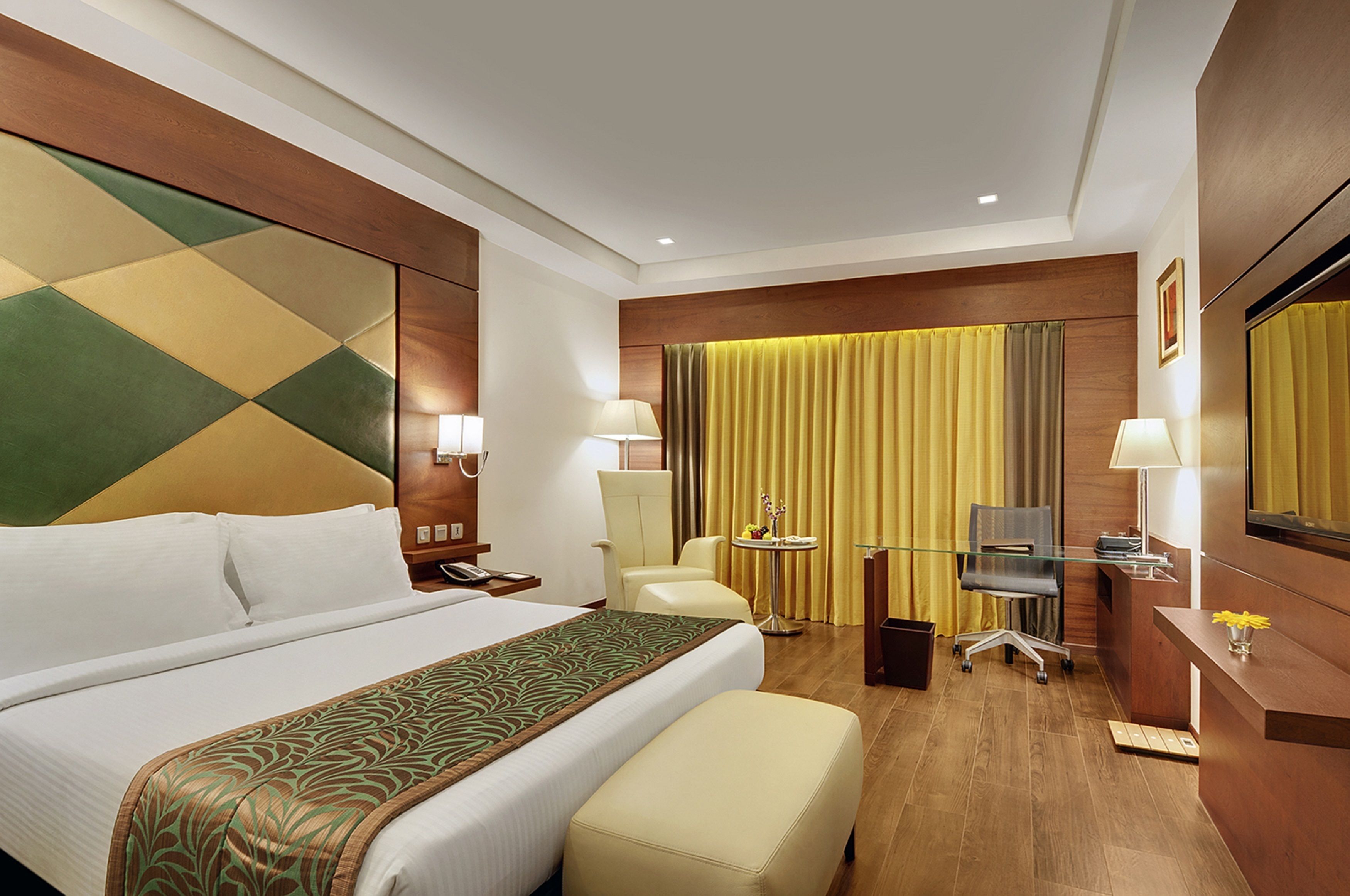 A hotel room with a large bed, a desk with a computer, a chair, and a small table with a lamp.