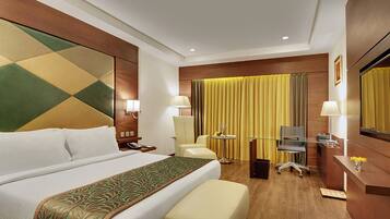 A hotel room with a large bed, a desk with a computer, a chair, and a small table with a lamp.