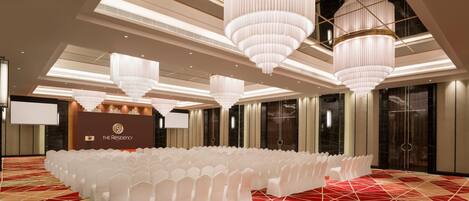A spacious conference room with rows of white chairs, large chandeliers, and a red patterned carpet.