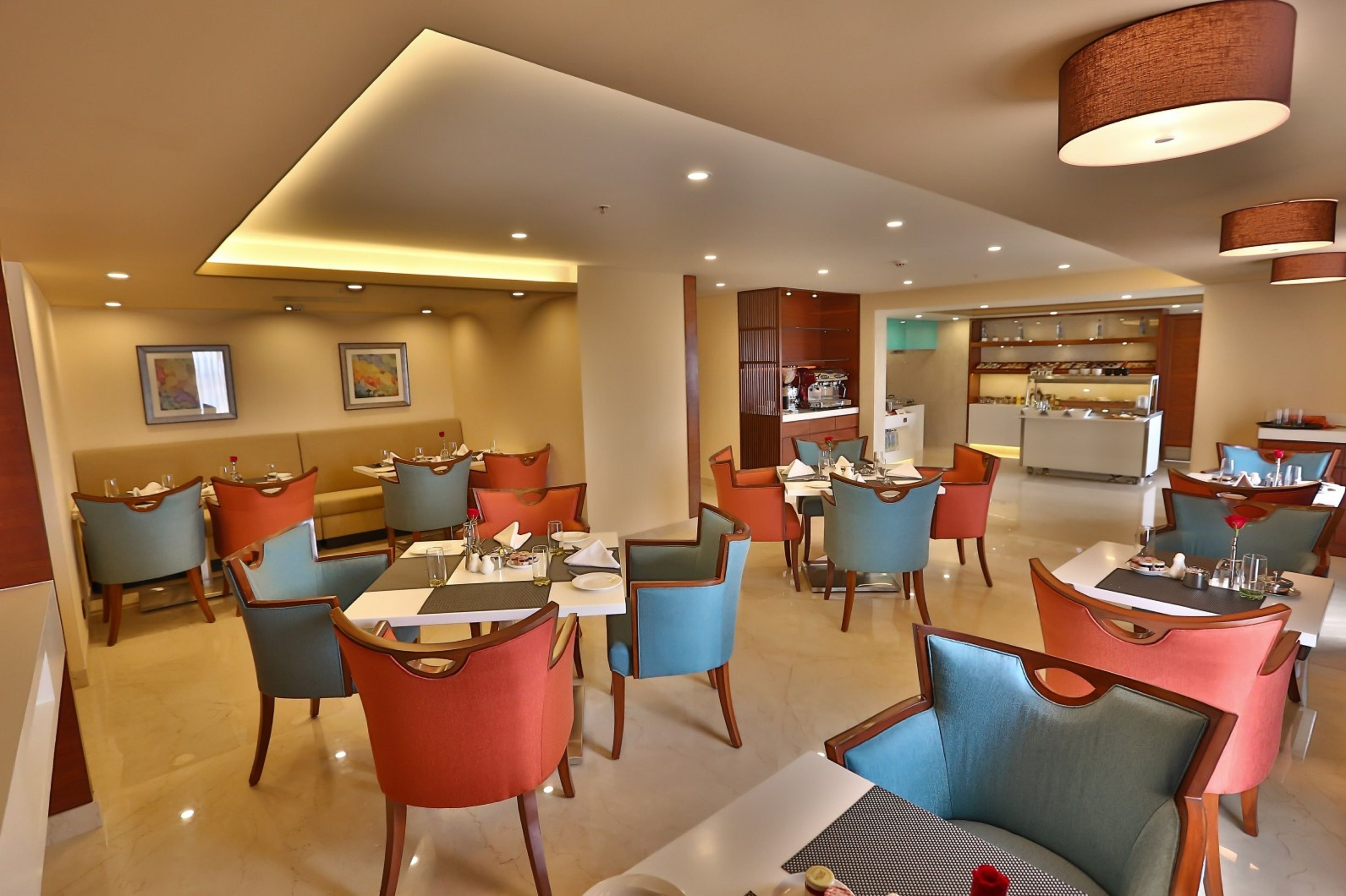 A modern dining area with multiple tables and chairs, a kitchen in the background, and a ceiling with recessed lighting.